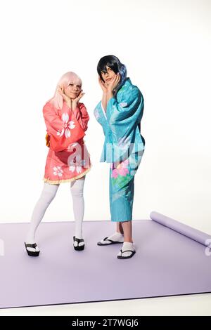 couple ludique de cosplayers dans une tenue vibrante touchant les visages sur tapis violet et fond blanc Banque D'Images