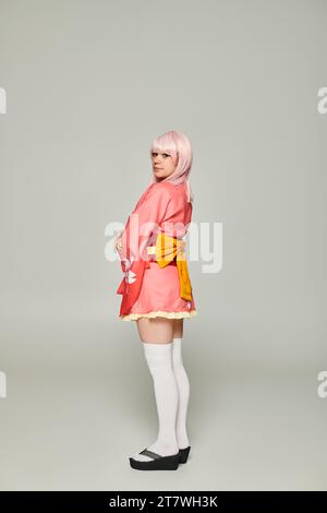 jeune femme de style anime en perruque blonde et kimono rose avec noeud jaune sur gris, concept cosplay Banque D'Images