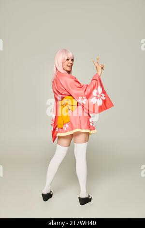 jeune femme en perruque blonde et kimono rose avec des chaussettes blanches au genou montrant le geste de victoire sur gris Banque D'Images