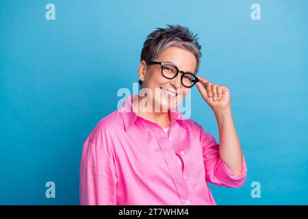 Photo de belle femme joyeuse intelligente avec des cheveux courts habillés doigts de chemise rose toucher des lunettes souriant isolé sur fond de couleur bleue Banque D'Images