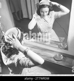 Femme devant un miroir dans les années 1940 Une jeune femme aux cheveux foncés dans une robe d'été rayée s'assoit devant le miroir et ajuste le chapeau assorti pour qu'il s'adapte parfaitement. Suède 1947. Kristoffersson réf. U122-5 Banque D'Images