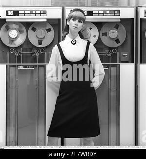 Dans les années 1960 Mannequin portant les vêtements typiques de 1965. La photographie est prise dans une salle informatique centrale des années 1960 avec des unités de bande magnétique utilisées où la bande contenant des informations est lue ou entrée. Elle porte une chemise blanche et une robe noire par-dessus. Photo prise 1965. Réf. DV16 Banque D'Images