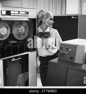 Dans les années 1960 Mannequin portant les vêtements typiques de 1965. La photographie est prise dans une salle informatique centrale des années 1960 avec des unités de bande magnétique utilisées où la bande contenant des informations est lue ou entrée. Elle porte une chemise blanche. Photo prise 1965. Réf. DV16 Banque D'Images