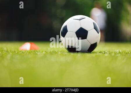 Ballon de football et marqueur à cône rouge sur le terrain d'entraînement. Gazon de football. Stade de football en arrière-plan flou. Jeunes joueurs pratiquant en arrière-plan Banque D'Images