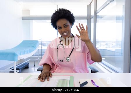 Portrait d'une femme médecin afro-américaine heureuse faisant un appel vidéo en agitant dans un bureau d'hôpital ensoleillé Banque D'Images