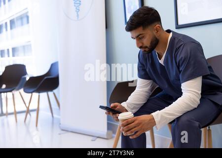 Médecin masculin biracial utilisant smartphone et boire du café dans la salle d'attente de l'hôpital Banque D'Images