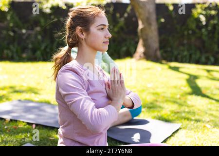 Femme biraciale focalisée pratiquant la méditation de yoga assise avec un ami dans le jardin ensoleillé, espace de copie Banque D'Images