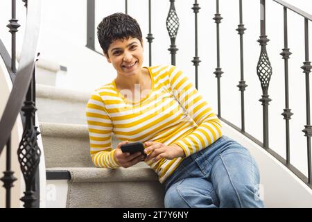 Portrait de femme biracial heureuse à l'aide d'un smartphone assis dans les escaliers à la maison Banque D'Images