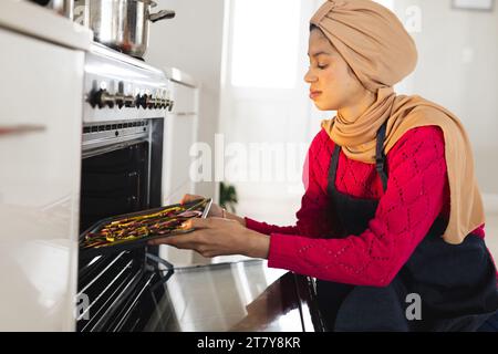 Femme biraciale en hijab et tablier mettant des légumes dans le four dans la cuisine Banque D'Images