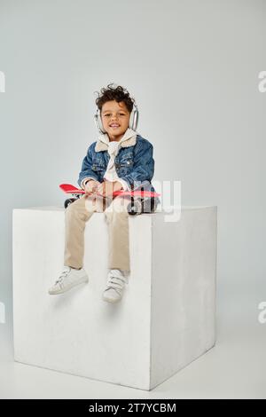 mignon garçon afro-américain en tenue d'hiver et écouteurs assis sur le cube avec penny board sur le gris Banque D'Images