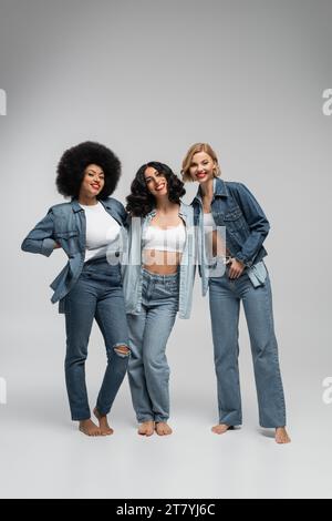 pleine longueur de copines multiraciales pieds nus dans des vêtements en denim bleu posant sur fond gris Banque D'Images