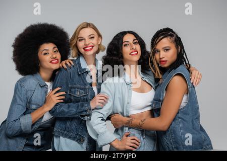 joyeuses copines multiethniques en vêtements denim tendance regardant la caméra tout en posant sur le gris Banque D'Images