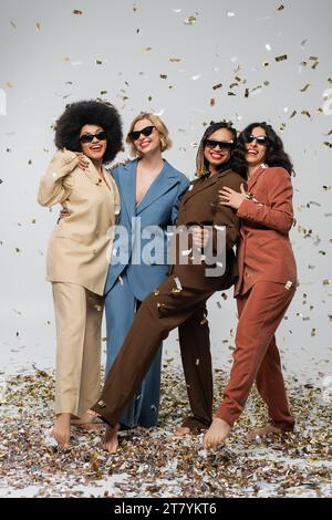 amies multiethniques insouciantes dans des lunettes de soleil et des costumes près de confettis colorés sur gris, temps de fête Banque D'Images