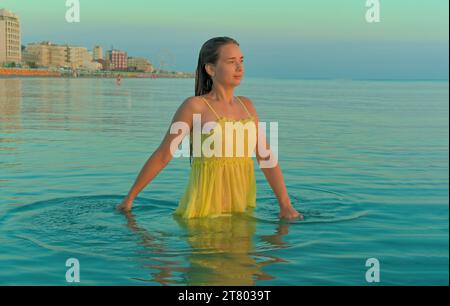 Une jeune femme va dans l'eau sur une plage de la ville tôt le matin en regardant le lever du soleil Banque D'Images