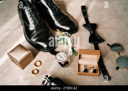 Les chaussures en cuir noir du marié reposent sur la table à côté des anneaux de mariage, boutonnière et montre Banque D'Images