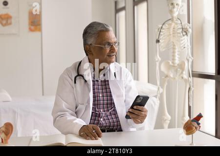 Praticien de médecine âgé réfléchi positif utilisant le téléphone portable Banque D'Images