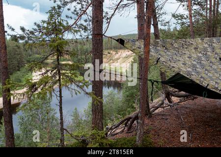 Un hamac avec bâche est installé parmi les arbres surplombant une rivière en contrebas dans la forêt sur un sentier de randonnée dans le nord de la Finlande Banque D'Images