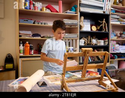 Beau garçon artiste debout près d'un bureau près d'un chevalet en bois, choisissant le croquis pour faire la reproduction d'image avec des aquarelles. Créativité, inspiration, art. Banque D'Images