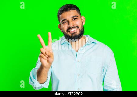 Heureux jeune homme indien montrant le signe de la victoire, espérant le succès et la victoire, faisant geste de paix, souriant avec une expression optimiste aimable, célébrer. Gars arabe isolé sur fond vert chroma clé Banque D'Images