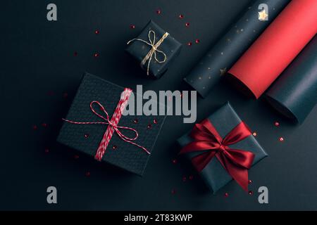Emballage de boîtes-cadeaux avec arc rouge et rouleaux de papier d'emballage sur fond noir. Noël festif, concept de célébration des vacances du nouvel an. Vue de dessus, Banque D'Images
