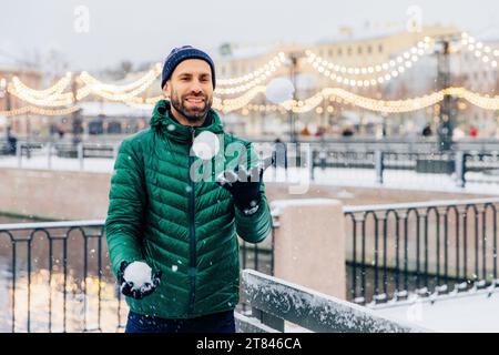 Homme profitant d'une journée enneigée, lançant une boule de neige, avec des lumières festives en arrière-plan Banque D'Images