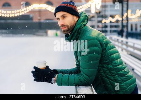 Homme contemplatif avec une tasse à café sur une patinoire enneigée, lumières festives au-dessus Banque D'Images