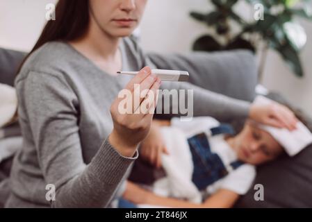 Focalisation sélective de la jeune mère traitant l'enfant fille malade souffrant de grippe et de fièvre, tenant le thermomètre, vérifiant la température corporelle élevée, appliquant Banque D'Images