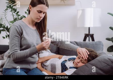 Inquiète jeune mère caucasienne traitant enfant fille malade souffrant de grippe et de fièvre, tenant le thermomètre, vérifiant la température corporelle élevée, appliquant Banque D'Images