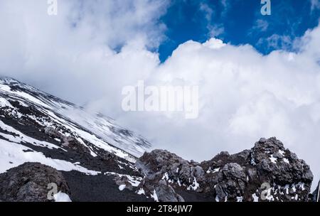 Pente du cratère de l'Etna, île de Sicile en Italie. Vue rapprochée des roches de lave volcaniques et des pierres couvertes de neige en hiver. Nuages et fumée de Banque D'Images