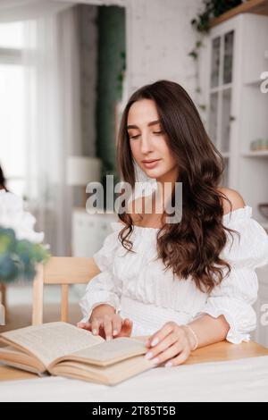 Séduisante jeune femme élégante avec de longs cheveux bouclés relaxant lit le livre de papier, retourne à travers une page, s'assoit à la chambre près de la table en bois pendant les loisirs Banque D'Images