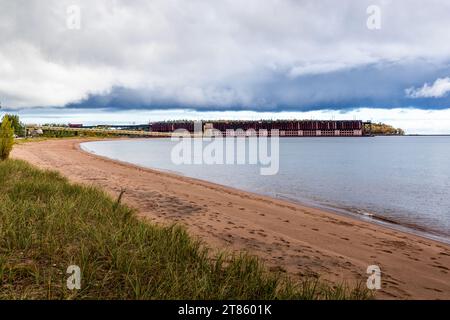 Le quai de Marquette Ore est en activité. Un cargo est amarré à la structure de chargement dans le port supérieur de Marquette, Michigan, États-Unis. Une ligne du Lake Superior & Ishpeming Railroad transporte du minerai de fer de la mine de Tilden jusqu'au quai, où il est chargé sur les bateaux de minerai du lac Supérieur. Construite vers 1911/1912, la structure est considérée comme l'un des derniers quais de minerai encore actifs dans le Michigan. Une plage de sable bordée de graminées dunaires s'étend en avant-plan. Banque D'Images