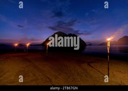 Les torches Tiki brûlent sur la belle plage tropicale et la silhouette des formations karstiques calcaires pittoresques s'élevant hors de la mer tropicale la nuit Banque D'Images