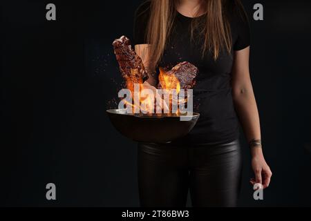 Femme aux mains tatouées, en leggings et t-shirt. Tenir la poêle wok avec le feu et frire deux steaks de bœuf sur fond noir. Gros plan Banque D'Images