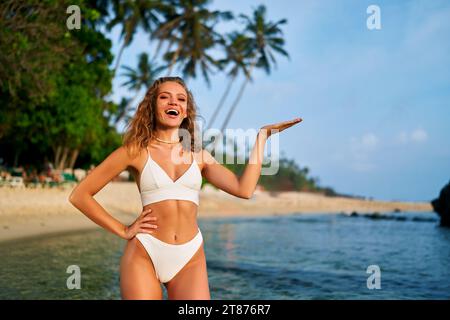 Femme joyeuse en bikini présentant avec la main sur la plage. Le modèle féminin souriant met en valeur le placement des produits à l'extérieur. Vêtements de plage d'été, tropical Banque D'Images