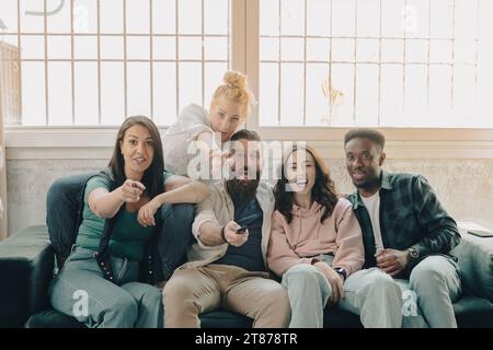 Fête à la maison parmi les jeunes. Groupe multiculturel d'enfants s'amusant à regarder la télévision sur le canapé. Rire et excitation devant le programme vu Banque D'Images