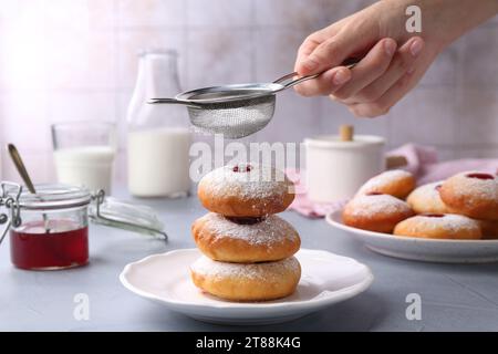 Femme saupoudrant le sucre en poudre sur de délicieux beignets Hanukkah sur la table gris clair, gros plan Banque D'Images