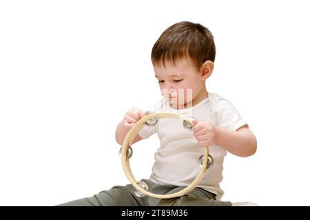 Bébé en bas âge joue du tambourin tout en étant assis sur le sol dans la chambre des enfants, isolé sur fond blanc. Enfant garçon jouant de l'instrument musical Banque D'Images
