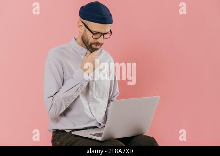 Photo de l'homme non rasé concentré est titulaire d'chin, regarde attentivement à l'écran d'ordinateur portable, bénéficie d communication en ligne, porte des lunettes et optique Banque D'Images