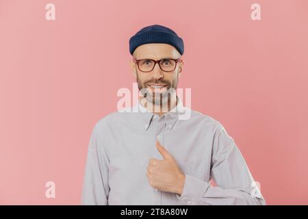 Satisfait l'homme a beau look attrayant, soulève le pouce, d'accord avec quelque chose, sourit joyeusement, porte un chapeau et chemise, plus isolé sur fond rose. Banque D'Images