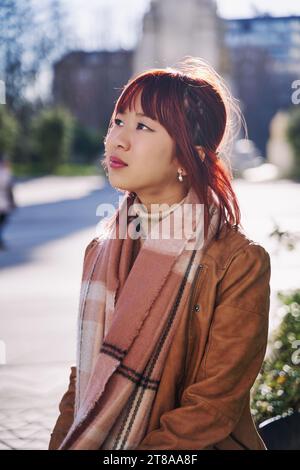 Jeune femme asiatique souriante confiante debout dans la rue, portrait. Fière femme portant une écharpe posant le regard loin dans la grande ville en plein air. Banque D'Images