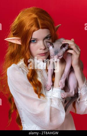 Portrait de cosplay elfe jeune femme en robe blanche, regardant attentivement à la caméra avec des yeux de différentes couleurs. L'elfe de Redhead tient le chat Sphinx endormi Banque D'Images
