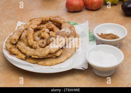 Une assiette blanche avec un motif de feuilles vertes autour du bord, contenant des torsions de pâte frite dorée fraîchement frite, avec deux petits bols de sucre en poudre Banque D'Images