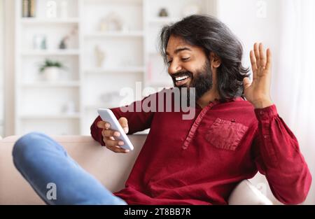 Homme indien positif assis sur le canapé et en utilisant smartphone Banque D'Images