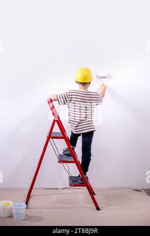 Un constructeur d'enfants se tient sur une échelle et peint les murs blancs d'un appartement. Un enfant avec un rouleau à peinture pendant la rénovation. Banque D'Images