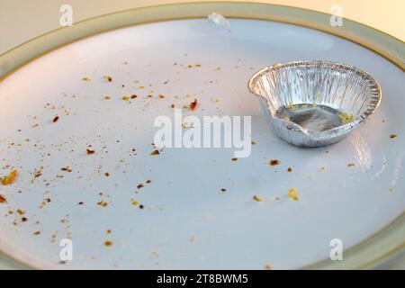 Une photo d'un moule à tarte en argent haché vide sur une assiette blanche recouverte de miettes sur un bureau blanc. Banque D'Images