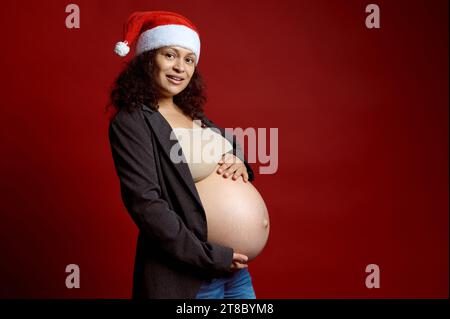 Charmante femme enceinte vêtue d'un élégant blazer gris et d'un chapeau de père Noël rouge, caressant doucement son gros ventre, souriant en regardant la caméra, isolé sur le rouge Banque D'Images