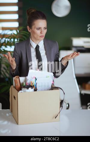 Nouveau travail. travailleur femme moderne stressé dans le bureau vert moderne en costume d'affaires gris avec des effets personnels dans la boîte en carton. Banque D'Images