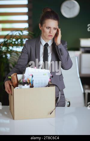 Nouveau travail. pensive femme moderne travailleur dans le bureau vert moderne en costume gris d'affaires avec des effets personnels dans la boîte en carton. Banque D'Images