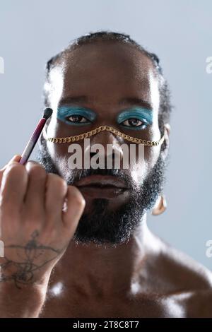 Homme gay noir confiant avec maquillage lumineux et accessoire. Debout isolé sur fond bleu clair. L'homme homosexuel afro-américain brosse le maquillage sur son visage. Cosmétiques et identité de genre Banque D'Images