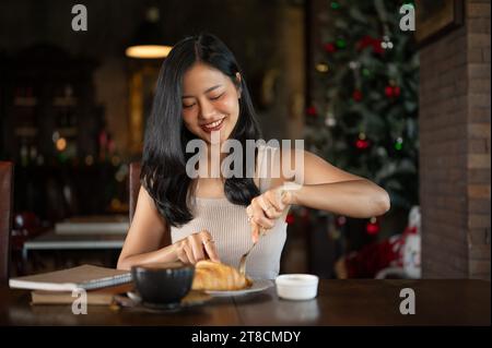 Élégante jeune femme asiatique mangeant un croissant pour le petit déjeuner avec latte chaud dans un salon de luxe sombre. Petit déjeuner du matin avec café. Banque D'Images